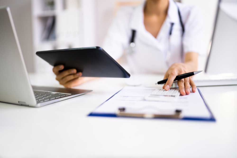 Doctor in white coat using tablet and writing at a desk with paperwork and laptop, representing medical billing tasks.
