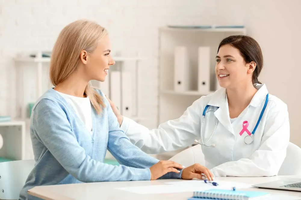 A doctor wearing a white coat and pink ribbon discusses thyroid cancer symptoms with a smiling patient in an office, raising awareness during Thyroid Cancer Awareness Month. | Cancer Biopsy & FNA Doctor in Los Angeles - Dr. Celina Nadelman