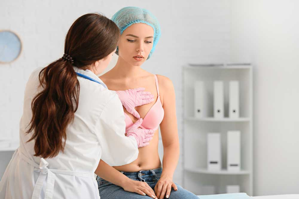 A doctor wearing gloves examines a woman’s breast during a medical appointment, as part of a comprehensive exam for thyroid cancer awareness month. The woman is seated, partially undressed, wearing a pink bra and a surgical cap. | Cancer Biopsy & FNA Doctor in Los Angeles – Dr. Celina Nadelman A doctor wearing gloves examines a woman's breast during a medical appointment, as part of a comprehensive exam for thyroid cancer awareness month. The woman is seated, partially undressed, wearing a pink bra and a surgical cap. | Cancer Biopsy & FNA Doctor in Los Angeles - Dr. Celina Nadelman